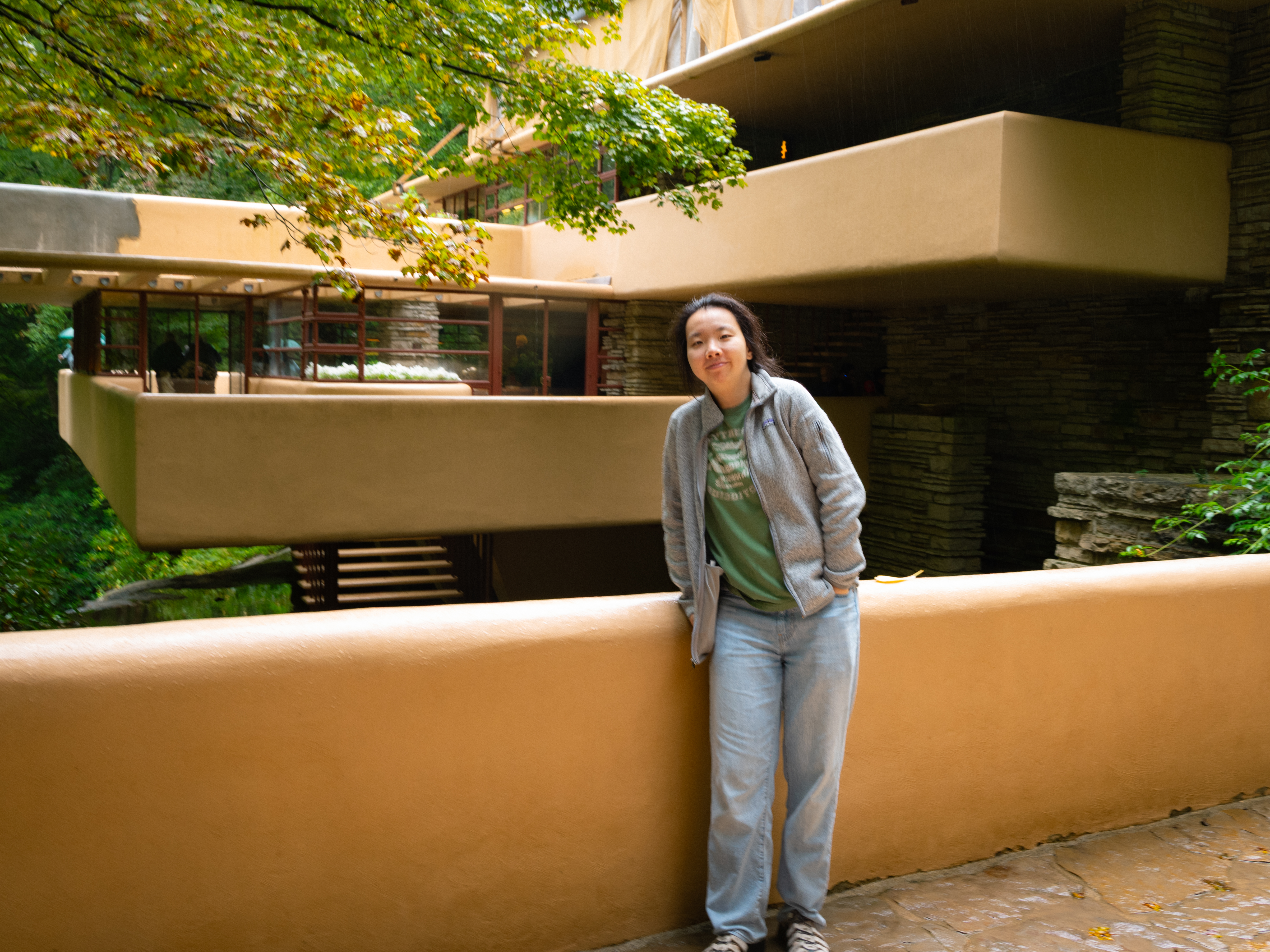 Zhixuan smiling shyly at Fallingwater, with Frank Lloyd Wright's cantilevered terraces and the surrounding forest in the background.